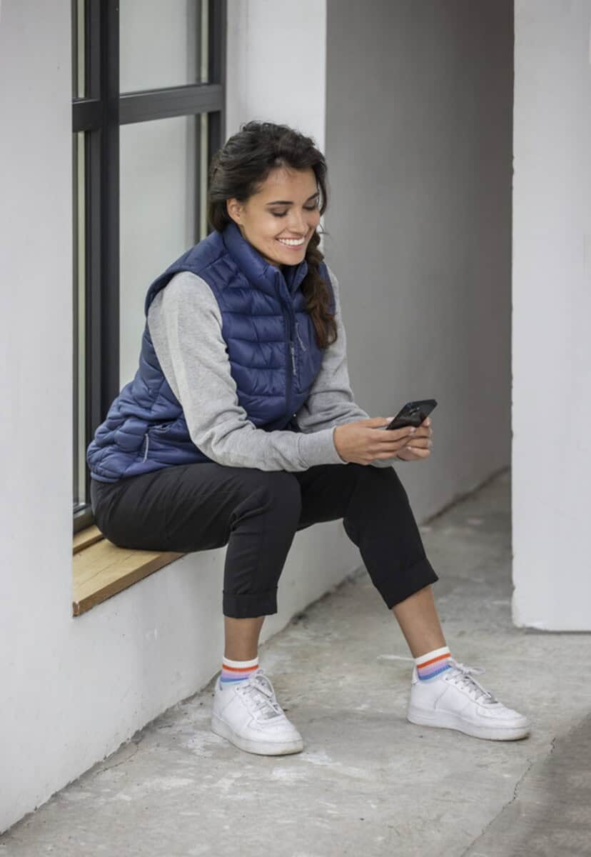 Bezrękawniki z logo – ocieplany bezrękawnik damski pallas z materiału z recyklingu 3943855_m1 1. Uśmiechnięta kobieta siedząca na parapecie, korzystająca z telefonu, casual styl, moda na co dzień, młodość, relaks, nowoczesny wygląd.